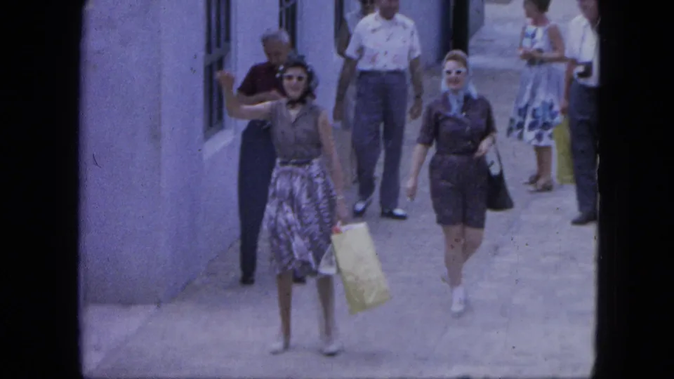 1964 Nassau street scene with adults in mid-century clothing walking beside a pale wall, including a woman waving and carrying a light bag.