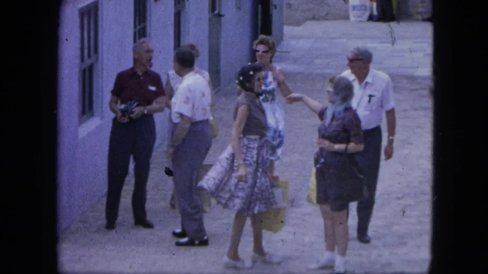A Nassau 1964 tourist group stands in a sunlit alley, with one man holding a camera and women carrying yellow shopping bags.