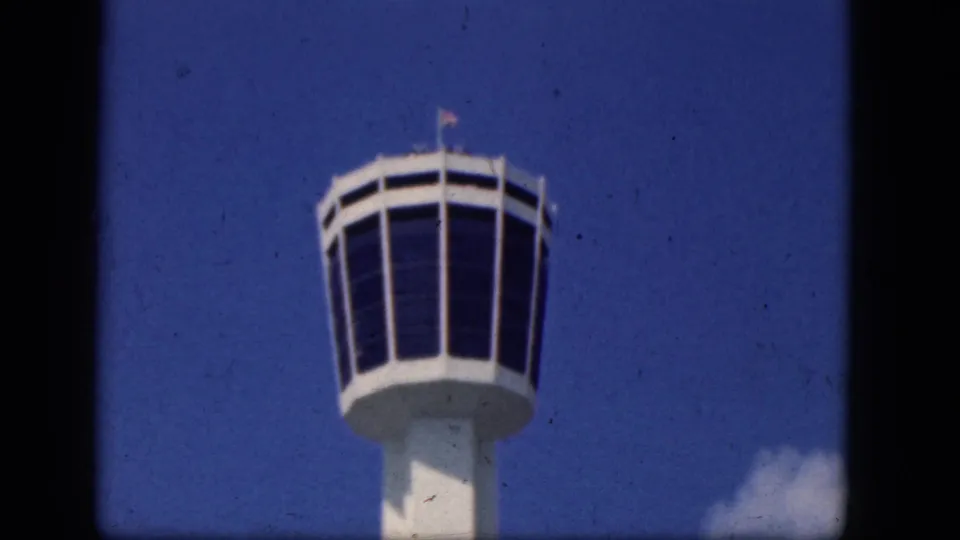 1964 8mm frame of a white airport control tower cab filmed from below in Niagra Falls, Canada, with blue sky behind it.