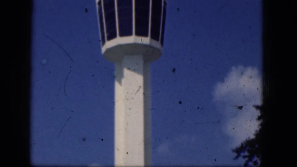 Ground-up 8mm view of the airport control tower, with its white column, dark windowed cab, and treetops at the lower edge.