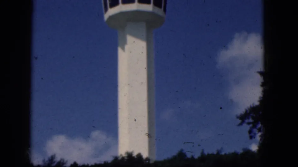 The control tower stands centered in a 1964 home-movie frame, with treetops below, clouds to the side, and visible film wear.