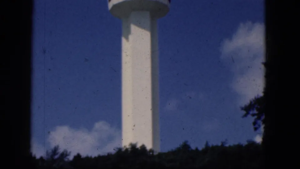 1964 8mm frame from Niagara Falls, Canada showing a white tower above a treeline, partly framed by dark foreground edges.