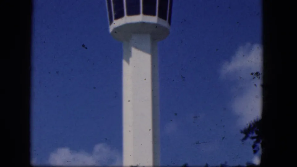1964 home-movie frame from Niagara Falls, Canada showing the upper shaft of a white airport control tower against blue sky.