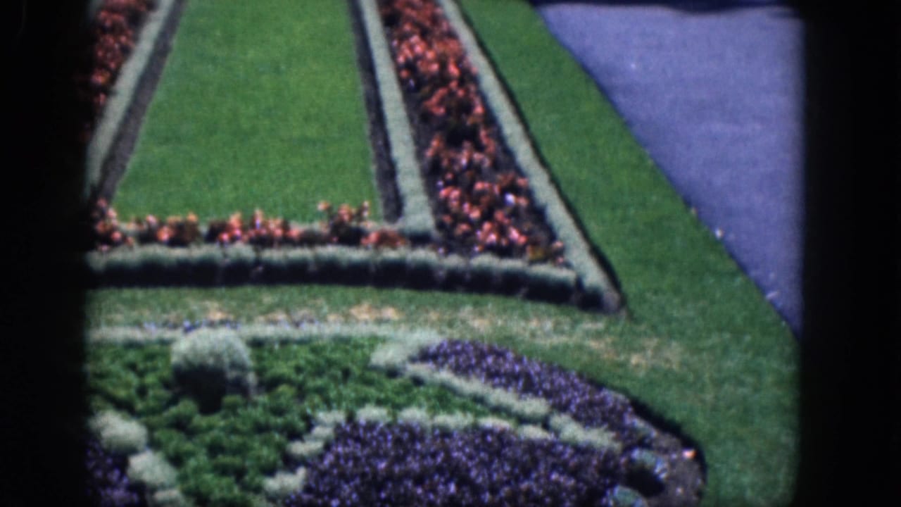 1964 8mm frame of Niagara Falls, Canada entrance gardens with red flowerbeds, trimmed grass, paths, and small distant visitors.