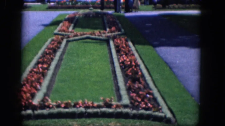 1964 8mm frame of Niagara Falls, Canada formal gardens showing red floral borders, lawn panels, hedges, and nearby pavement.