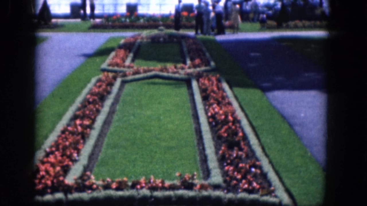 Formal flower beds and grass lead toward a waterfall overlook with visitors in the distance.