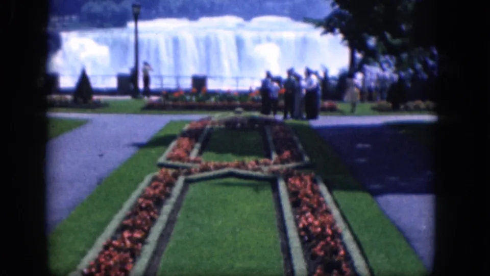 Mid-century 8mm travel frame of a Niagara Falls, Canada garden, with clipped lawns, red flowers, visitors, and the falls in back.