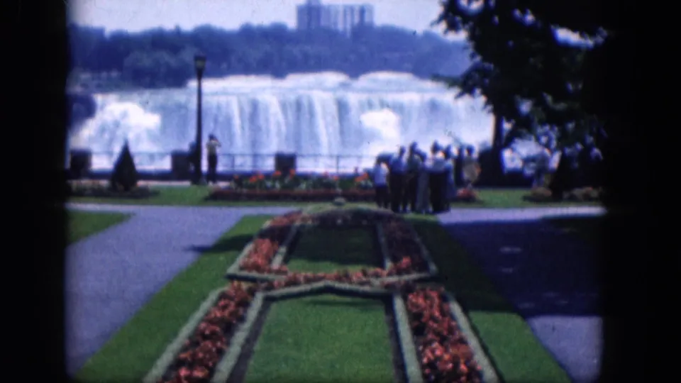 Wider 1964 Niagara Falls home-movie frame with shaded trees, formal flower beds, sightseers, railings, and the waterfall beyond.