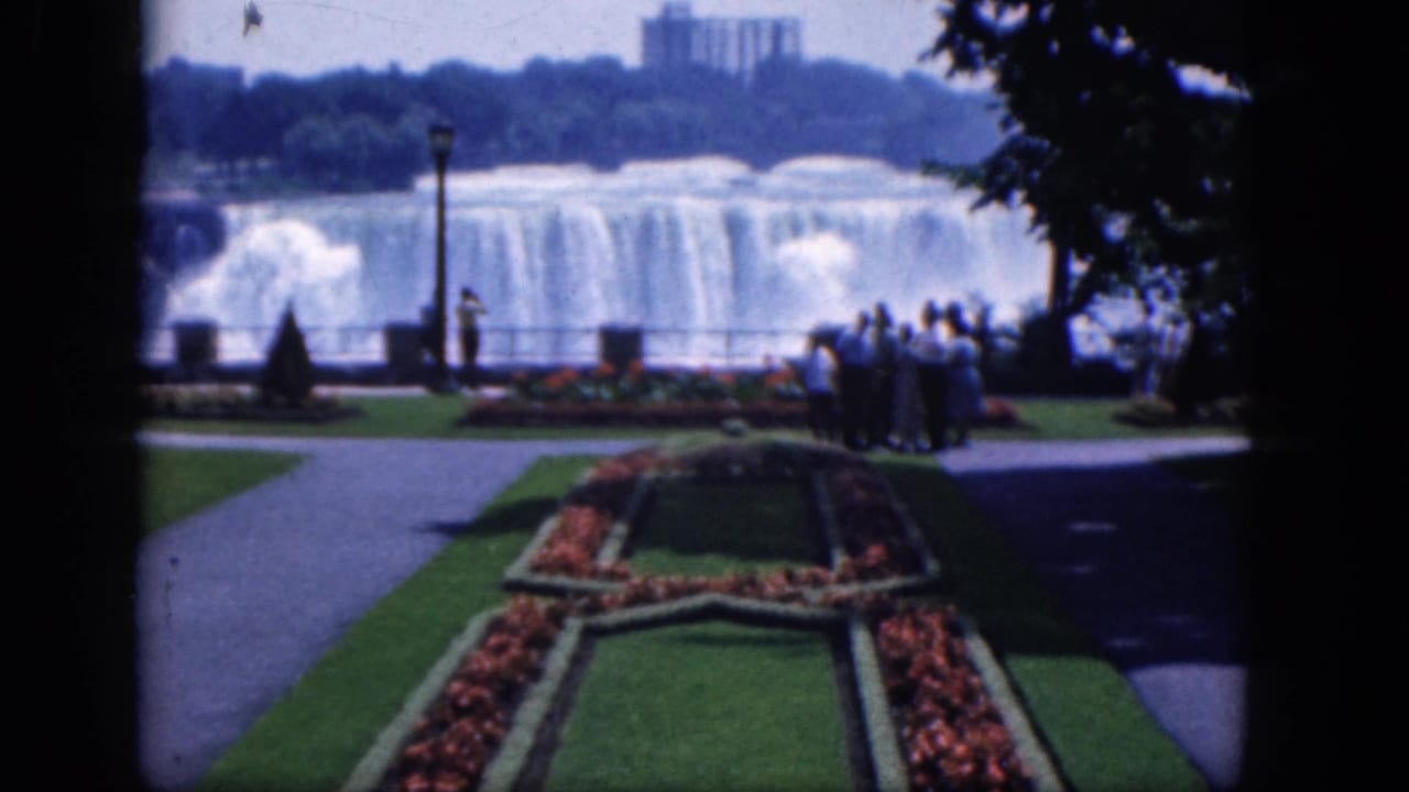 Tourists view Niagara Falls from a landscaped overlook with paths and flower beds.