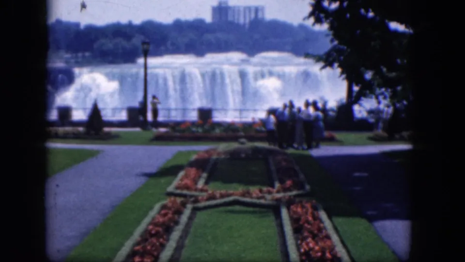 1964 8mm home-movie view of tourists standing near a railing at Niagara Falls, Canada, framed by lawns, paths, flowers, and trees.