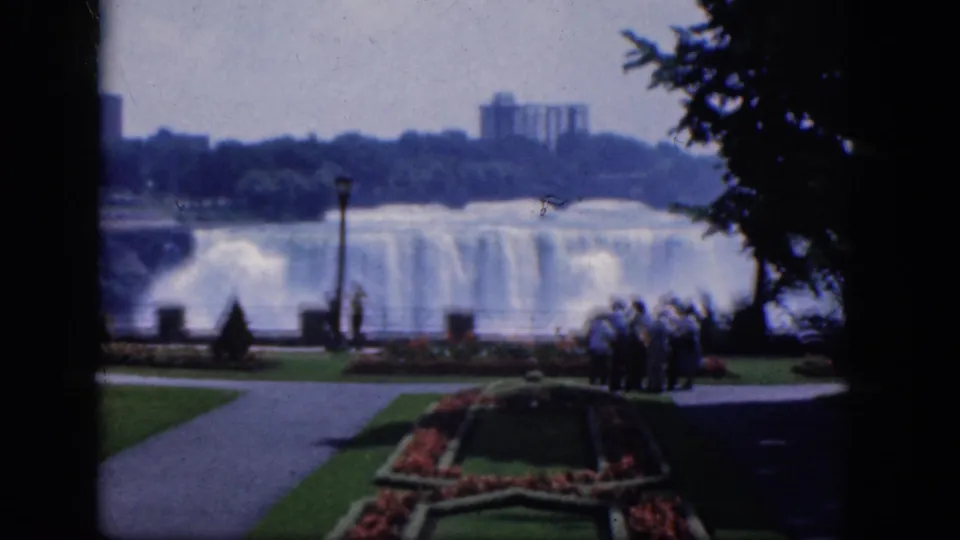 A handheld 8mm frame shows Niagara Falls behind tourists at a landscaped Canadian overlook, with paths, lawn, flowers, and trees in view.
