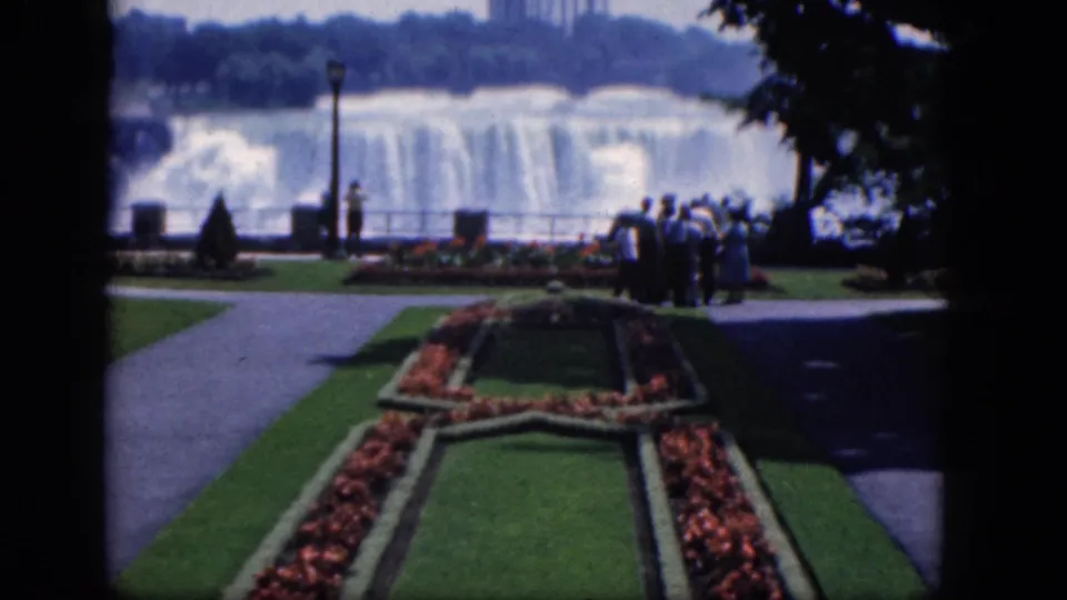 The 1964 home-movie frame places tourists at the right of a landscaped overlook, with Niagara Falls, walkways, flower beds, and shaded trees visible.