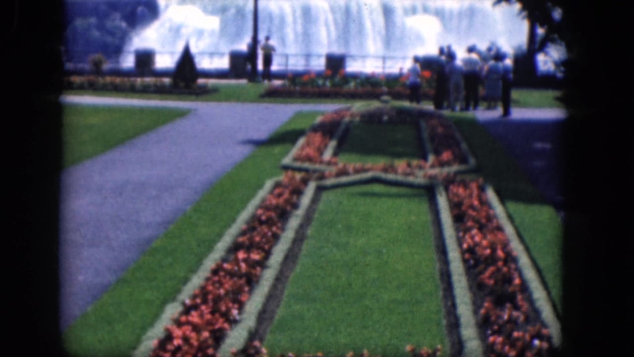1964 Niagara Falls, Canada view across a manicured garden toward tourists standing near the waterfall railing.