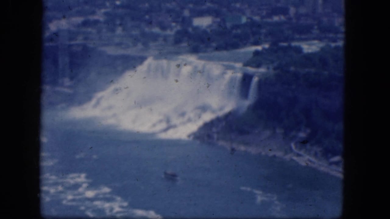 Waterfall and river seen from above, with mist, shoreline, distant buildings, and dark film borders.
