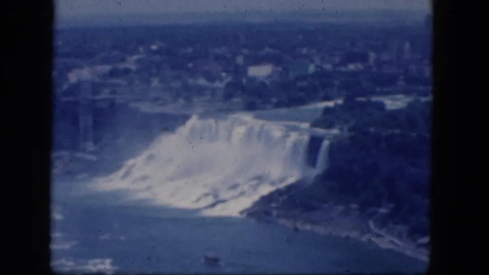 1964 8mm frame showing Niagara Falls from an elevated angle, with broad river water, white mist, shoreline, trees, and distant development.