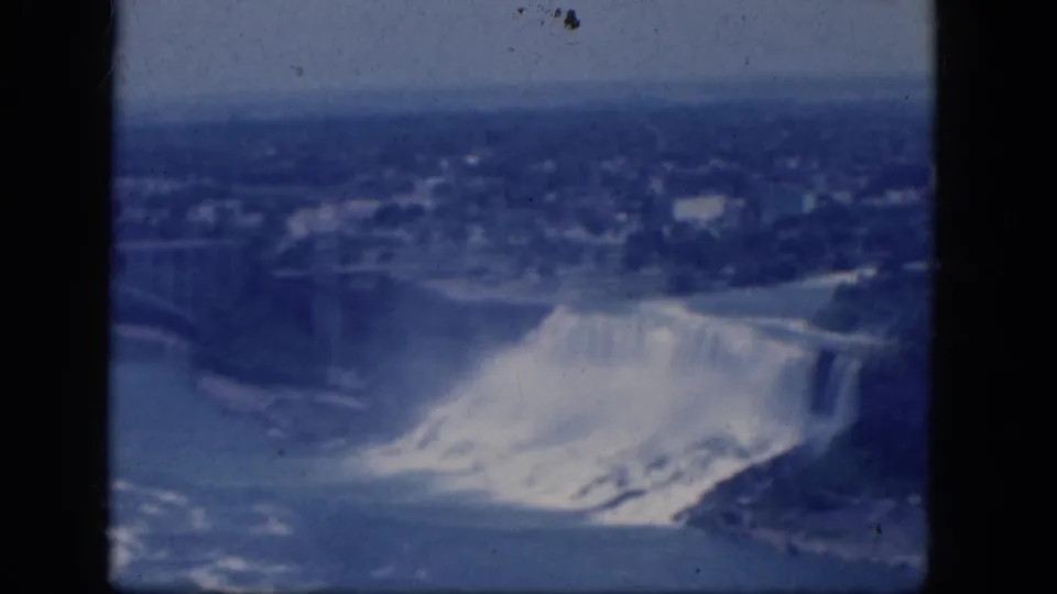 1964 Niagara Falls 8mm frame with the waterfall, river, mist plume, shoreline, nearby trees, and distant landscape under a blue faded cast.