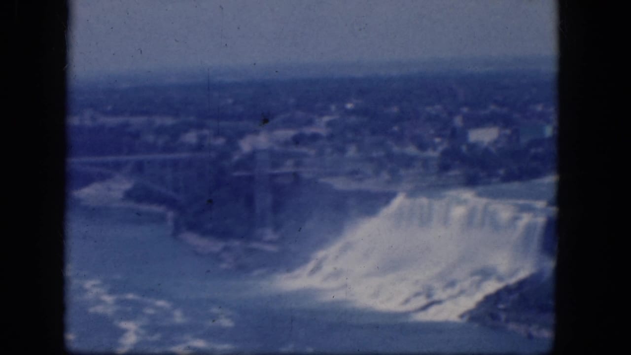 1964 8mm view of Niagara Falls from an elevated vantage, with a bridge and hazy shoreline beyond the waterfall.