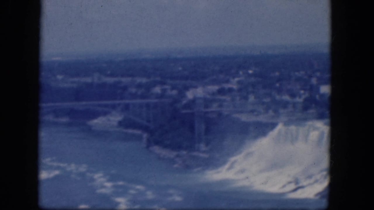 Blue-tinted 1964 home-movie frame showing Niagara Falls, the river and a bridge from a high viewpoint.