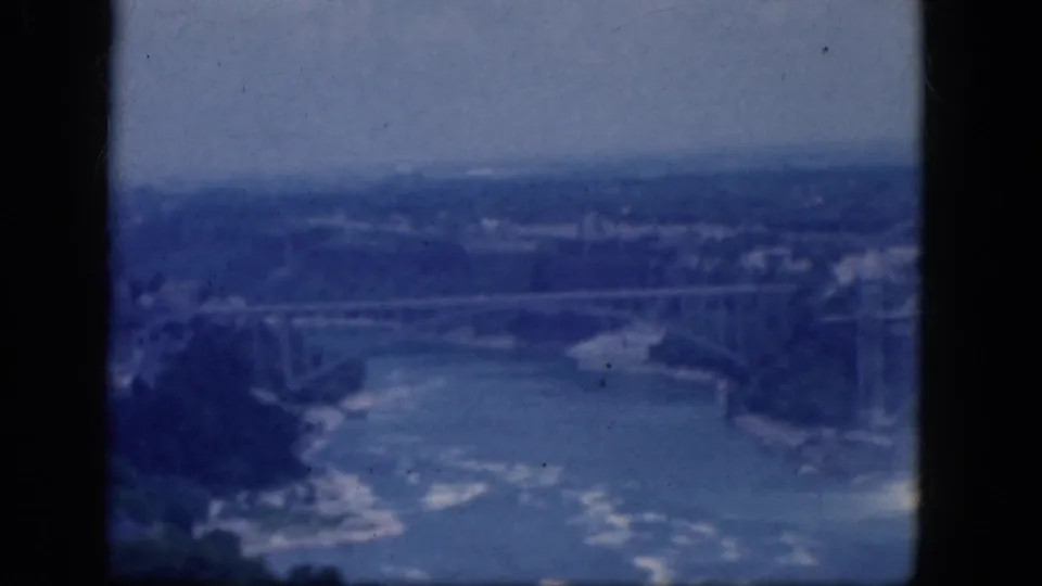 Soft blue 1964 8mm frame looking across the Niagara River toward a bridge, with waterfall spray and shoreline visible.