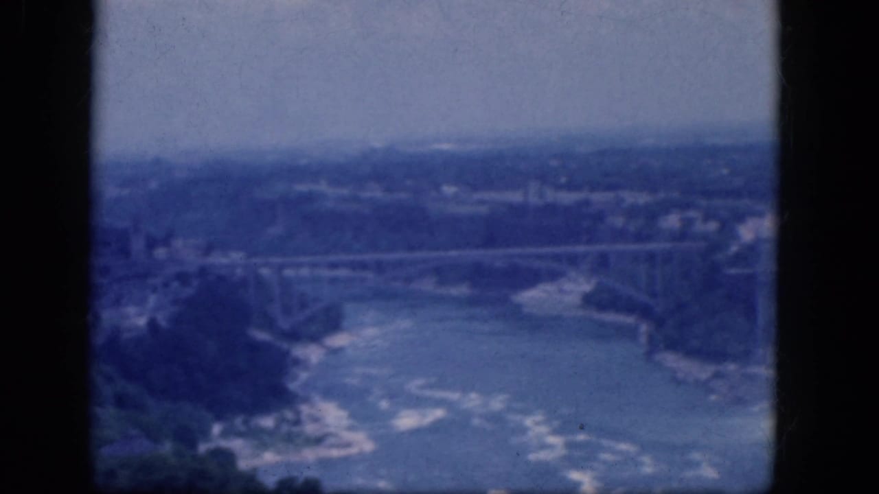 Soft 8mm view of Niagara Falls area with river, gorge, bridge, and distant townscape.