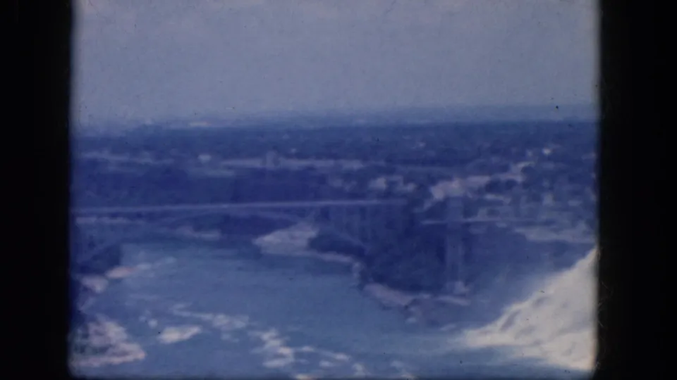 1964 Niagara Falls home-movie frame showing the falls at right, a bridge over the gorge, and distant buildings beyond the river.