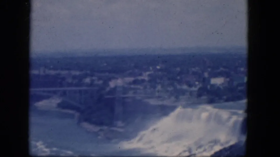 1964 home-movie view from above Niagara Falls, with cascading water, river bend, bridge structure, and distant townscape.