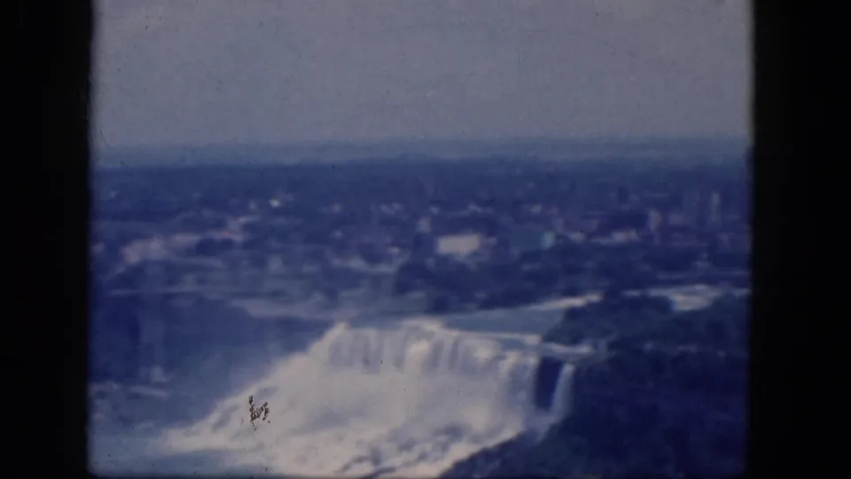 1964 8mm frame of Niagara Falls from a high overlook, showing misting water, the river, shoreline, and distant urban buildings.