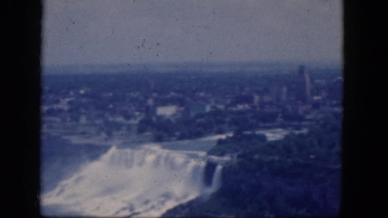 Aerial view of Niagara Falls with mist, river water, trees, and a distant skyline.