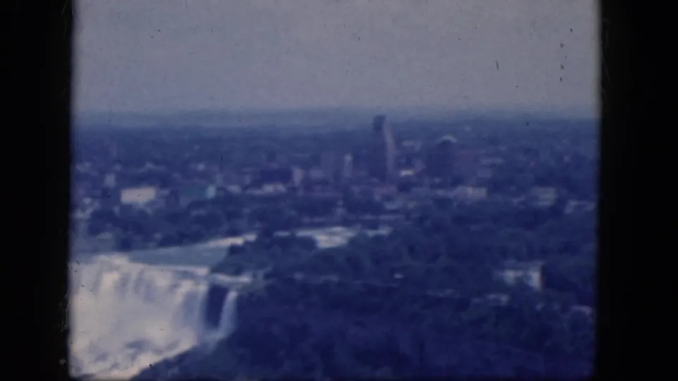 Aerial 8mm frame of Niagara Falls, Canada, showing the falls at left, wooded parkland, haze, and distant city buildings.