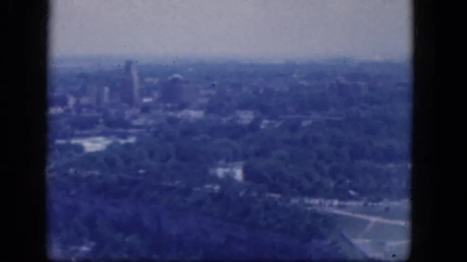 1964 Niagara Falls, Canada aerial view with distant high-rise buildings, dense trees, and soft blue home-movie exposure.