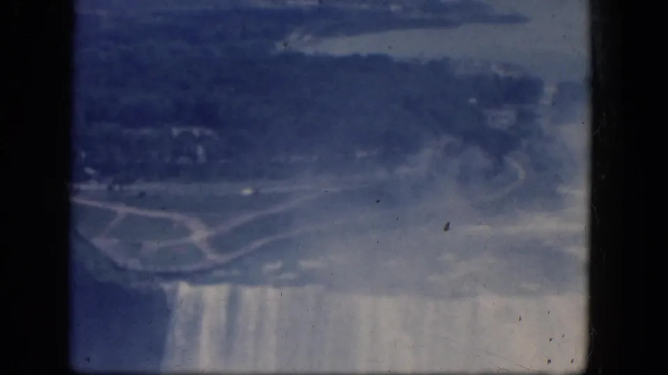 1964 8mm frame showing the Horseshoe Falls edge from above, with heavy mist, cascading water, and nearby shoreline paths.