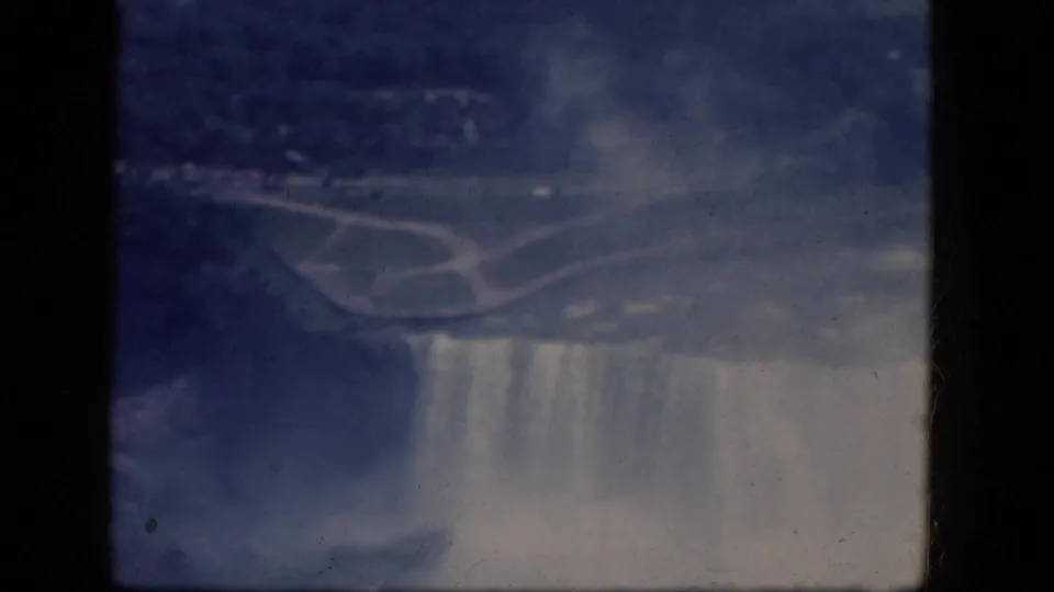 1964 Canadian-side Niagara Falls frame showing the curved Horseshoe Falls brink, mist, shoreline, and a distant road.