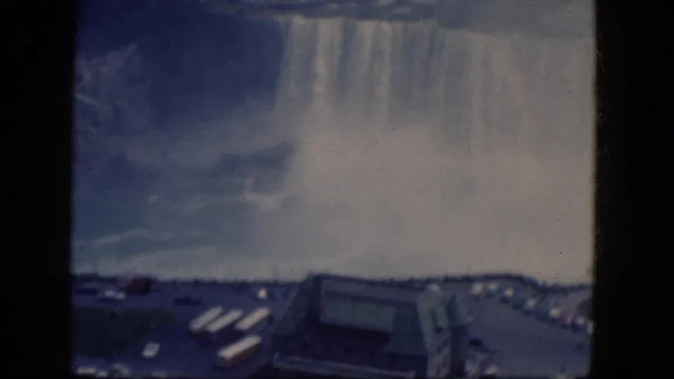 1964 Niagara Falls home-movie frame showing mist above the falls and a parking area with cars and a large visitor building below.