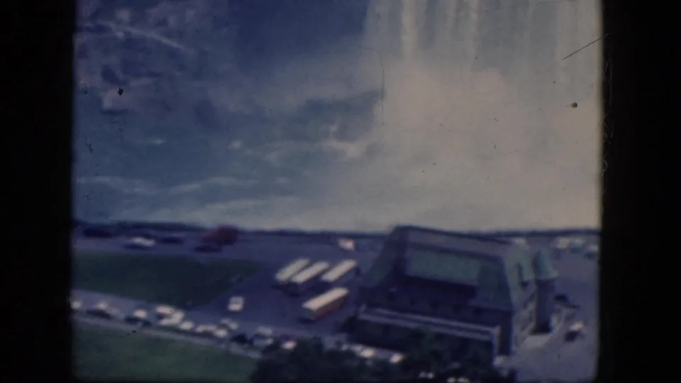 Canadian-side Niagara Falls view with mist, roads, parked cars, tour buses, and a large visitor building in a 1964 8mm frame.