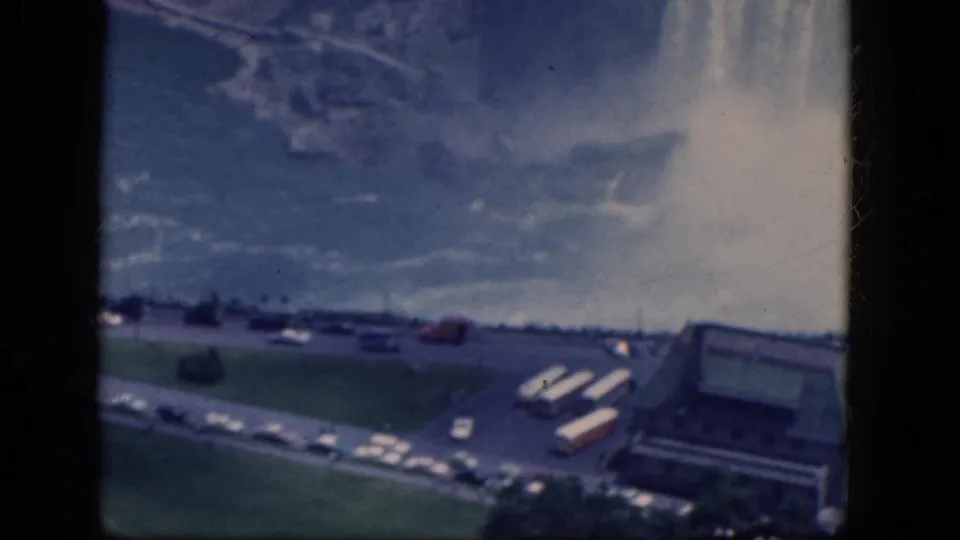 1964 8mm aerial view near Niagara Falls, Canada, with mist rising behind roads, parked cars, tour buses, lawns, and a visitor-area building.
