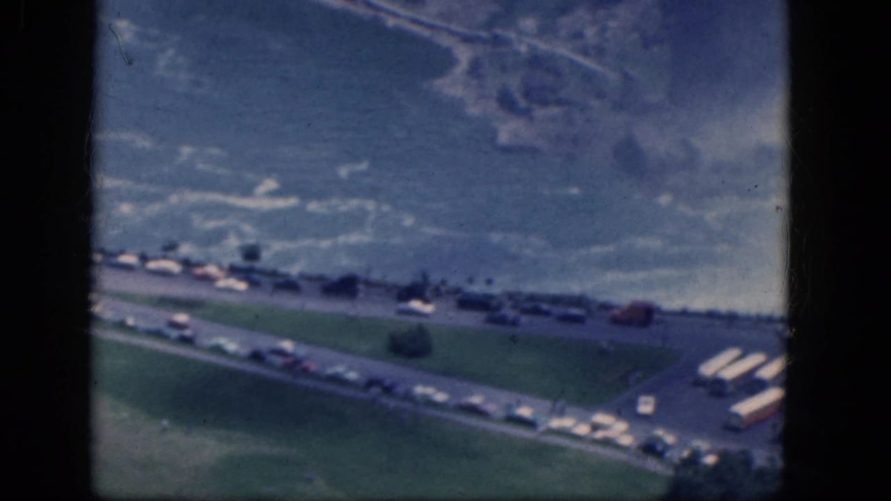 Aerial view of mist, roadway, green lawn, parked cars, buses, and distant buildings.