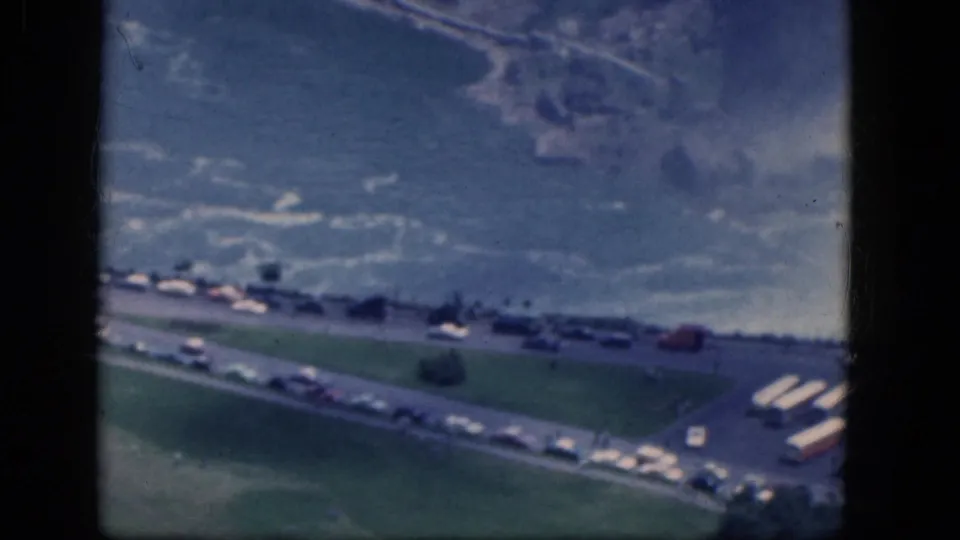 1964 Niagara Falls aerial frame showing waterfall mist beyond a road, green lawn, parked cars, tour buses, and distant visitor-area buildings.