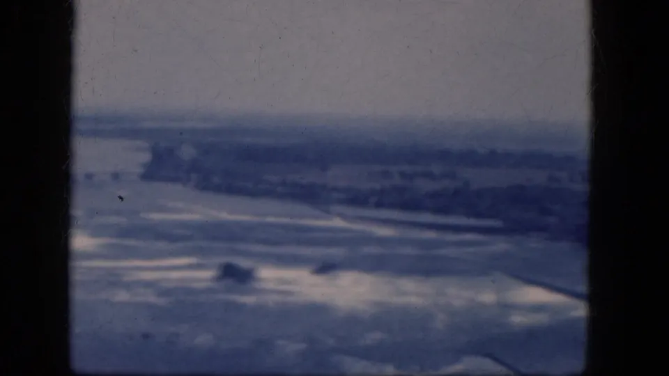 Blue-tinted 1964 home-movie aerial view with distant land and waterlike forms below the aircraft window.