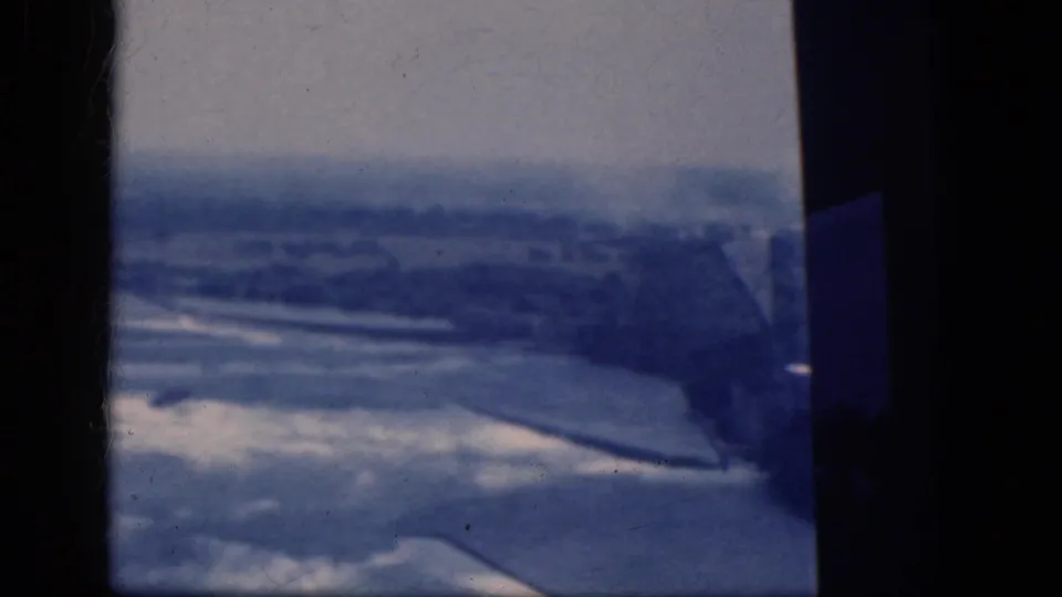 Soft 8mm aerial frame from the supplied 1964 Niagara Falls, Canada clip, showing indistinct terrain below an aircraft.