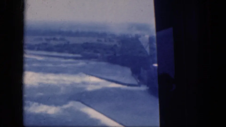 Blue 8mm aerial view from the 1964 Canada travel clip, with distant structures and pale terrain below the window.
