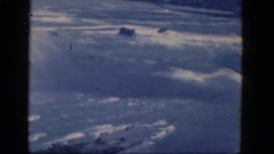The 8mm frame looks past dark aircraft window divisions toward a blue aerial landscape with pale ridges and darker patches.