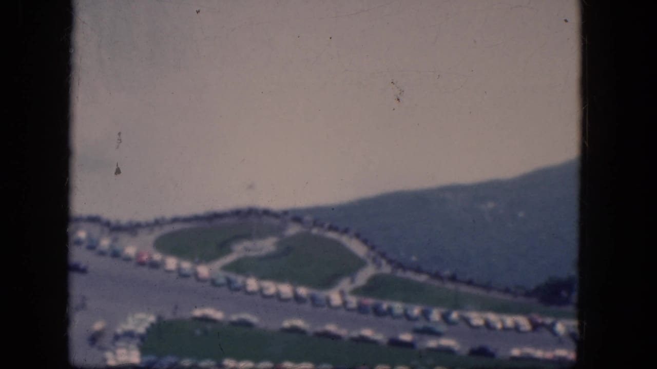 Elevated view of a winding road, parked cars, lawns, and a distant hillside.