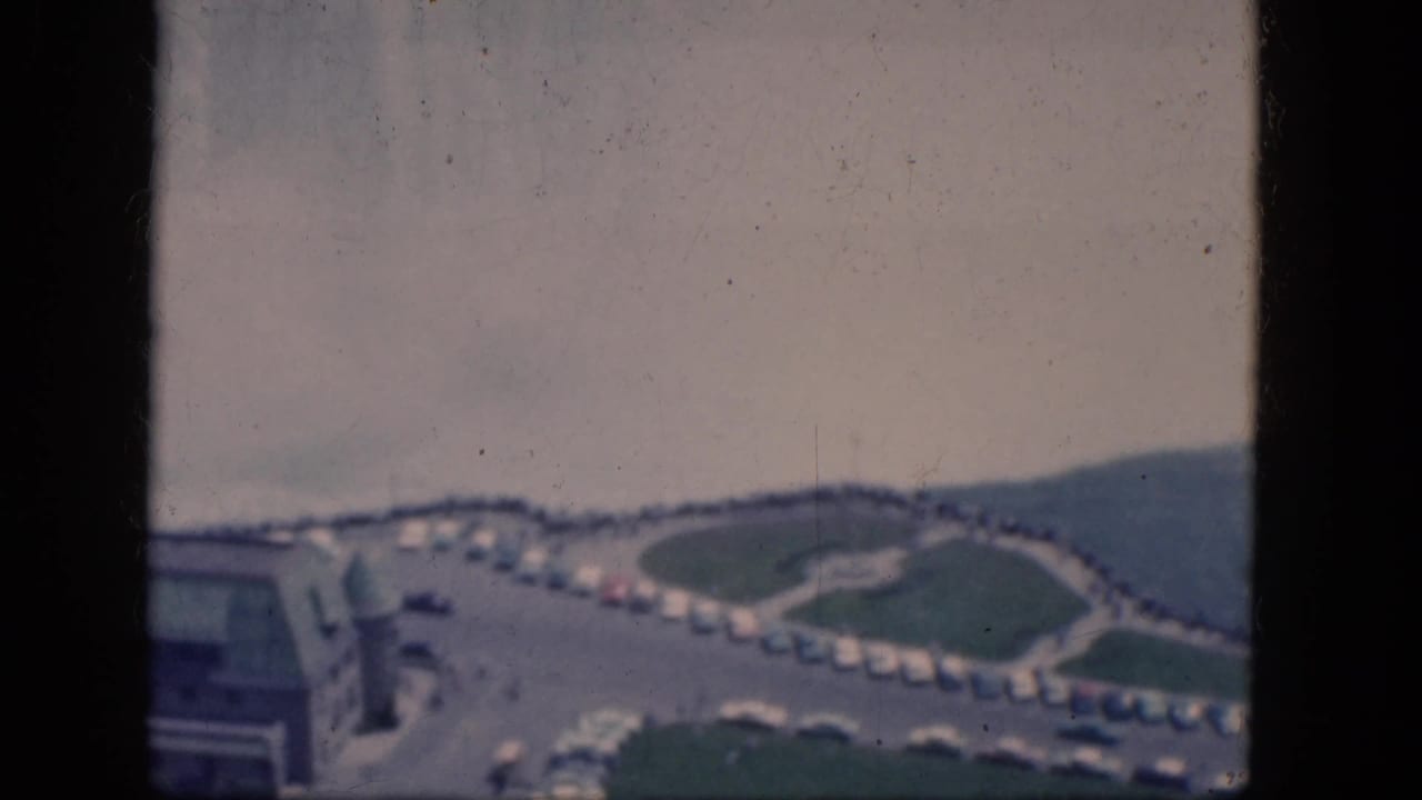 Curving roadway with parked cars, lawns, and a hillside in the background.