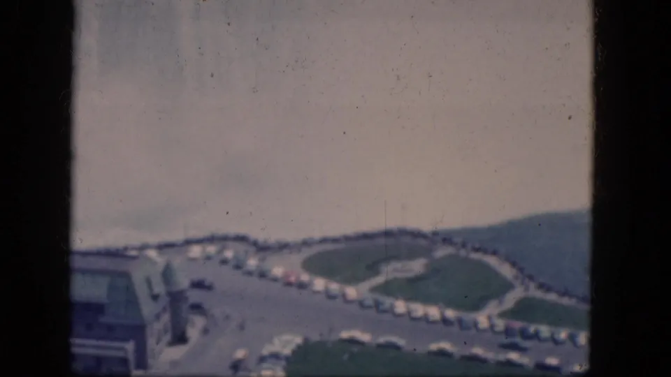 1964 Niagara Falls, Canada travel footage frame with a curved road, rows of parked cars, green lawns, and a distant hill.
