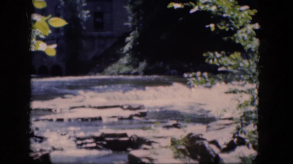 1964 Niagara Falls, Canada travel frame showing flowing water, scattered rocks, foliage, and dark stonework in soft focus.