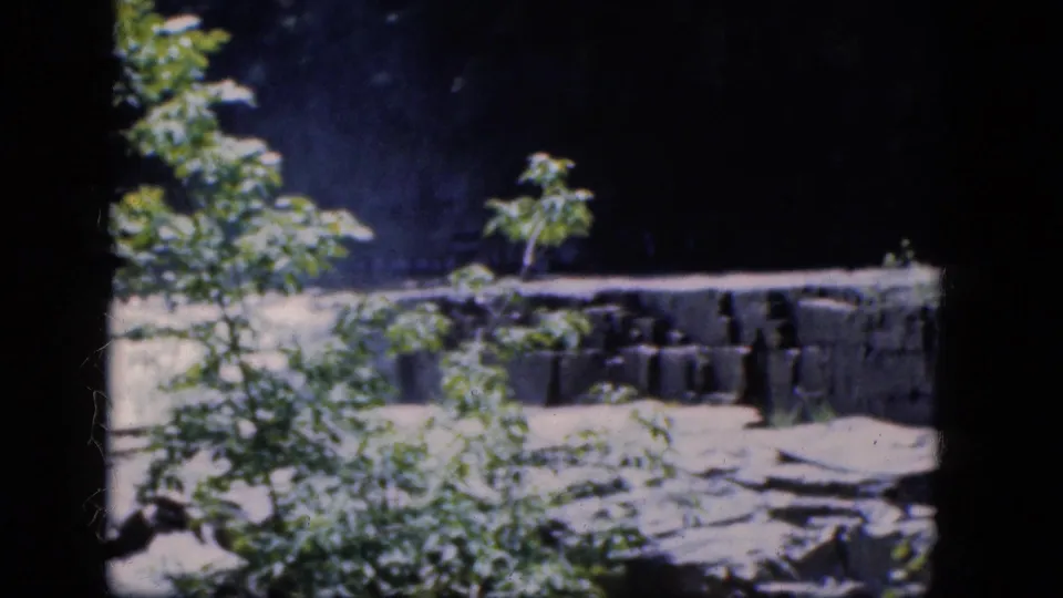 1964 Niagara Falls home-movie frame showing a low stone retaining wall near creekside rocks and foliage.