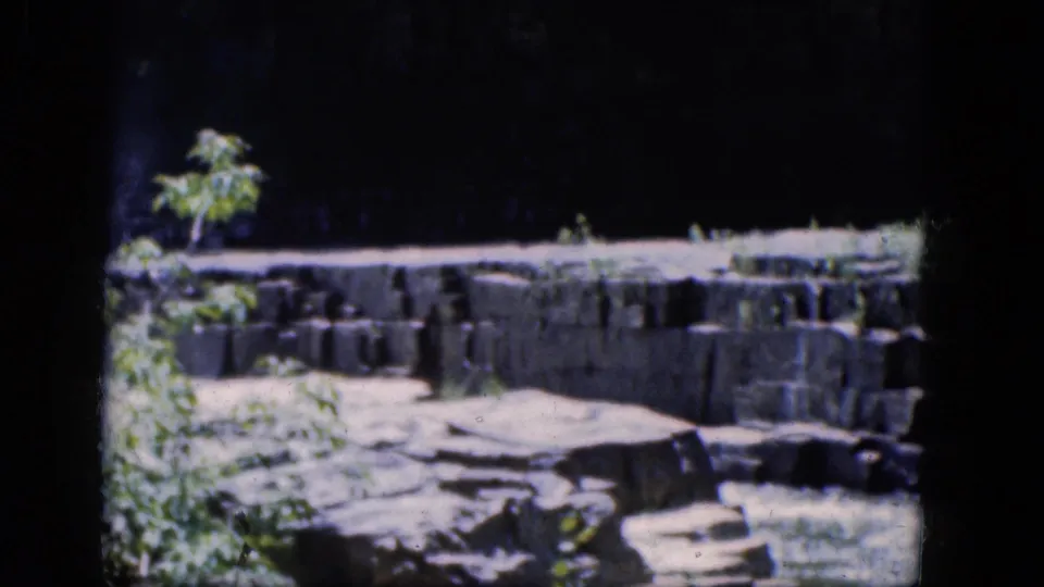 1964 8mm frame from Niagara Falls, Canada showing a rocky creek edge, stone wall, and plants under strong shadow.