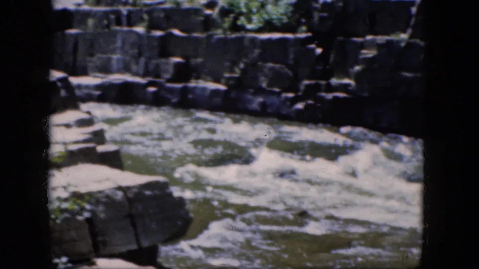 1964 home-movie frame showing swift Niagara Falls river water below dark rock ledges and sunlit stone shelves.