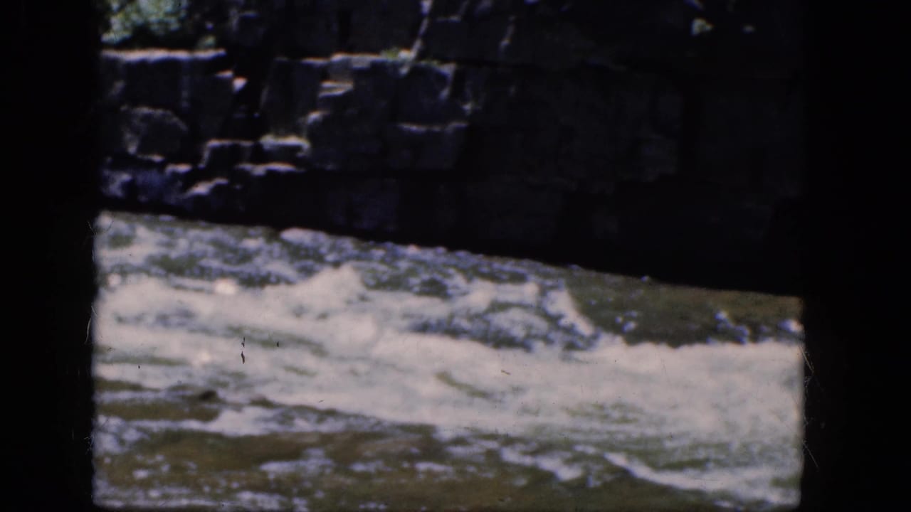 Foaming river water rushes below a stone retaining wall with dark film borders.