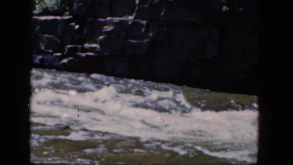 1964 8mm footage from Niagara Falls, Canada shows churning white water below a weathered stone wall, framed by dark film edges.
